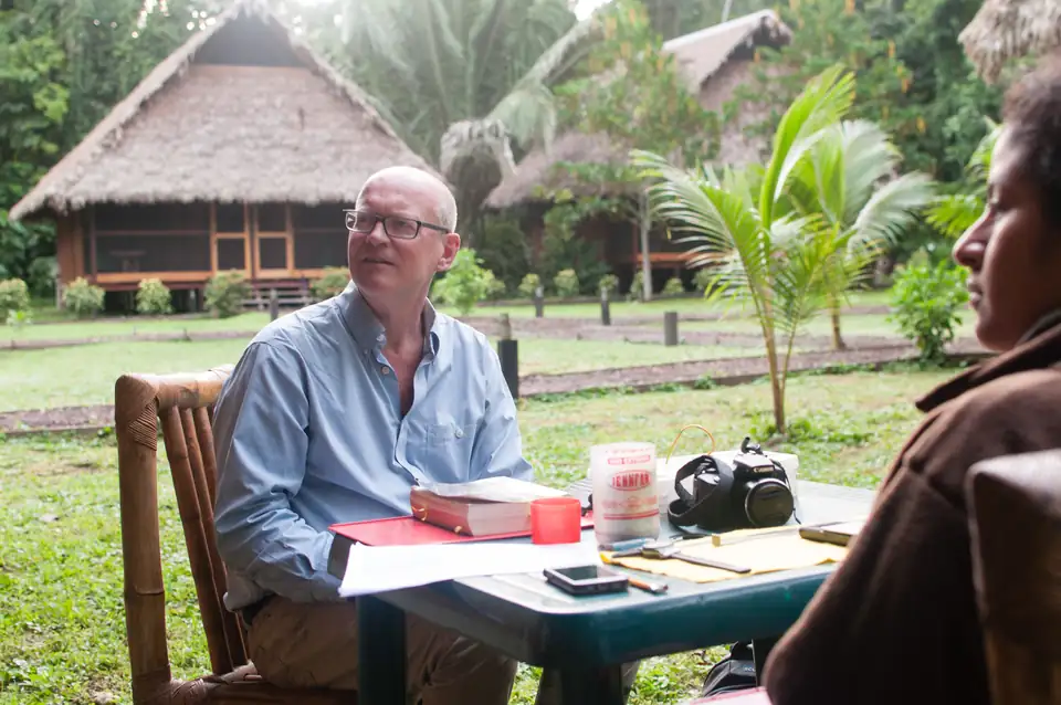 A researcher records bird data at a table in front of a thatched building